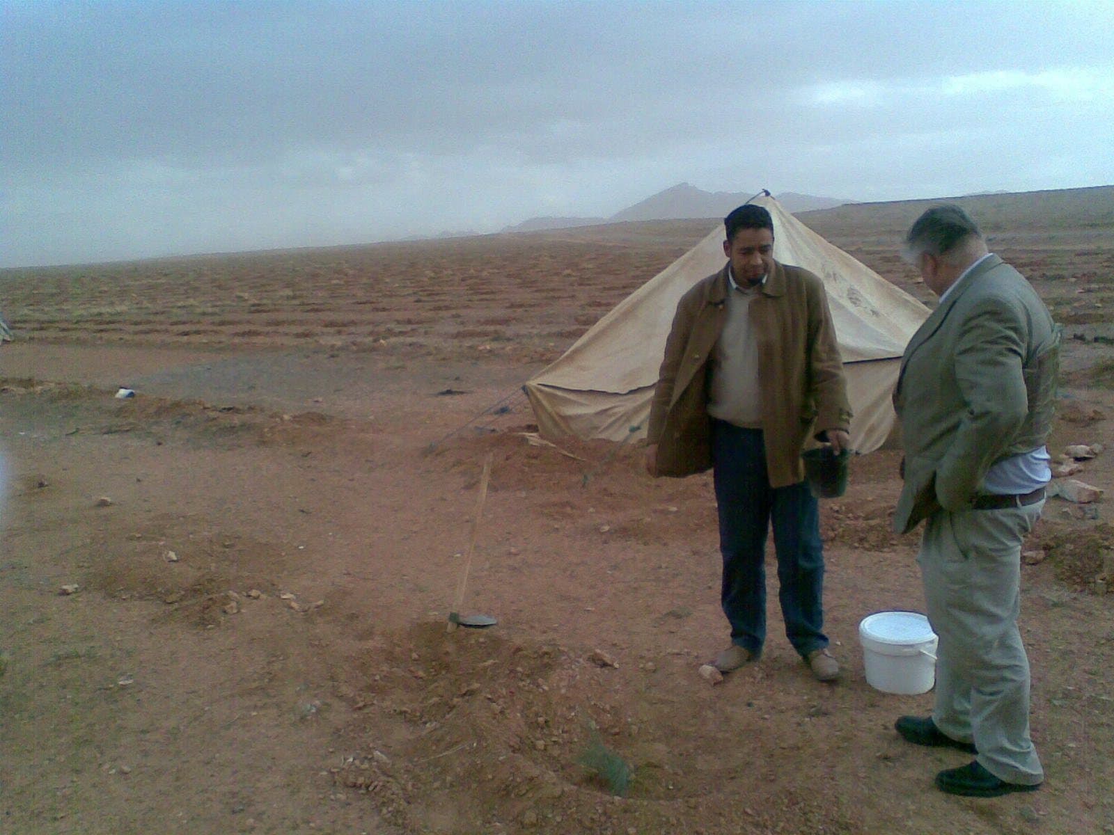 Two men standing beside a canvas tent on flat, stony desert ground at Bouarfa under a grey sky